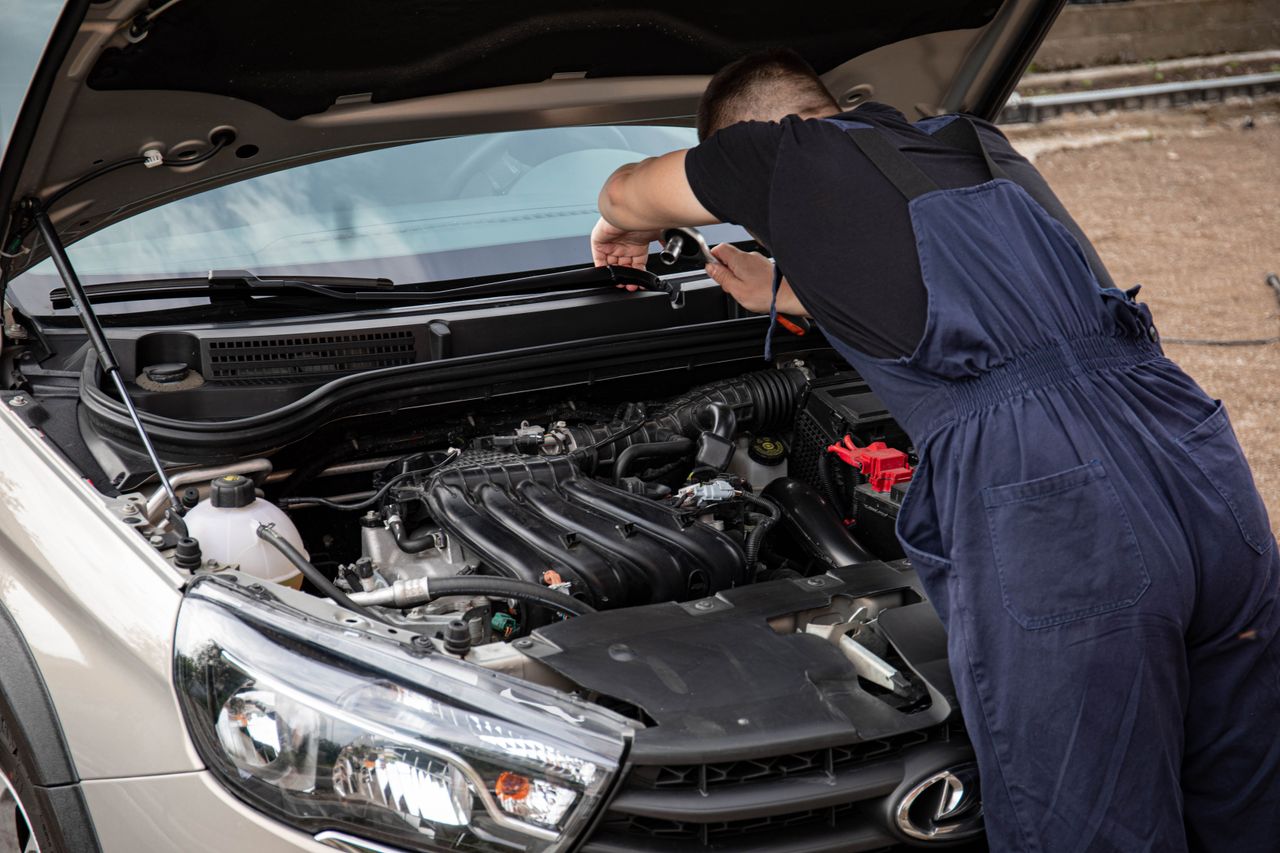 A mechanic in blue overalls works on a car engine in an outdoor setting.